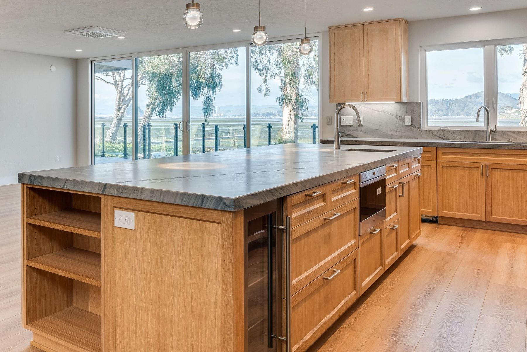 Kitchen island of light wood and stone opening to a bay through sliding glass.