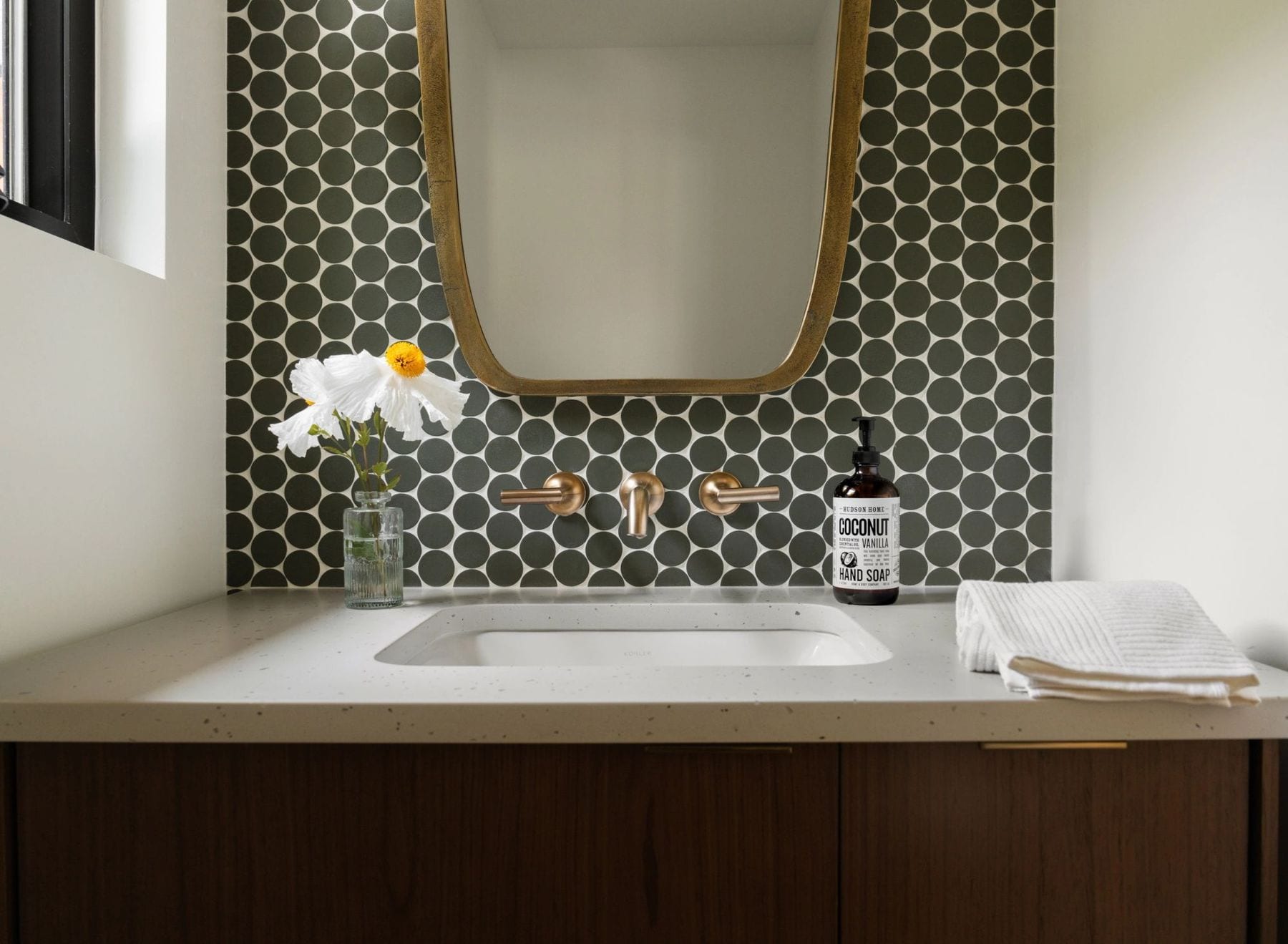 Bathroom vanity with wood cabinets, gold fixtures, and a dark penny-tile backsplash.